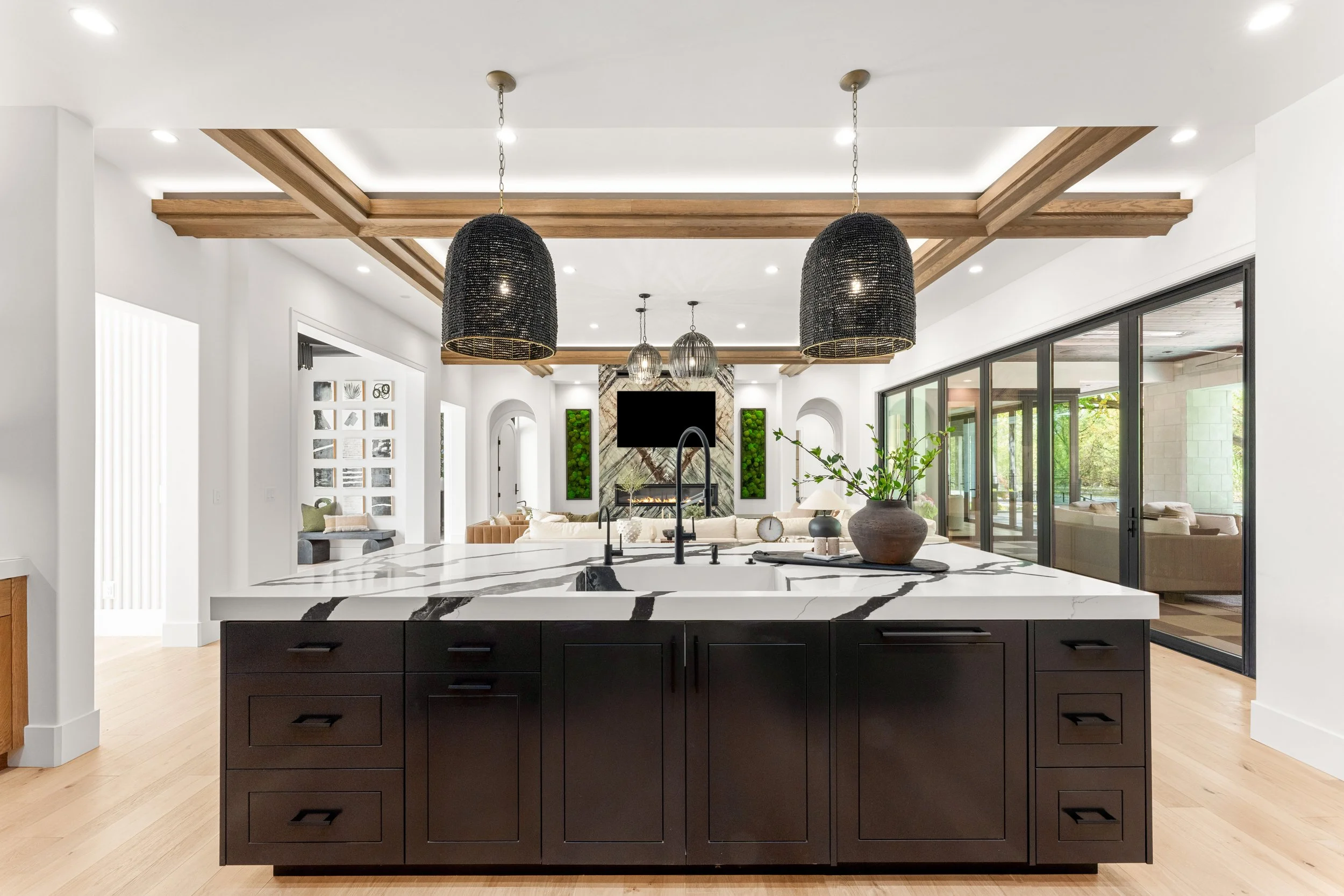 Modern open-concept living space with a black kitchen island, marble countertops, black pendant lights, wooden ceiling beams, large windows, and a fireplace with a TV above.