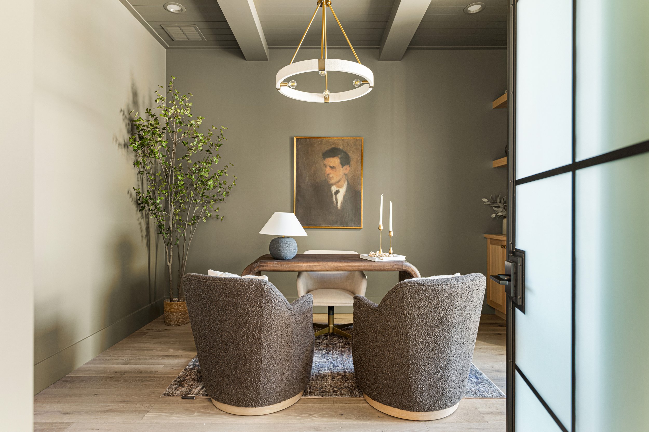A cozy dining area with two upholstered chairs around a wooden table, a large green potted plant, a painting of a man, a white table lamp, and candles with decor on a tray, with a modern chandelier overhead and a glass-paneled room divider.