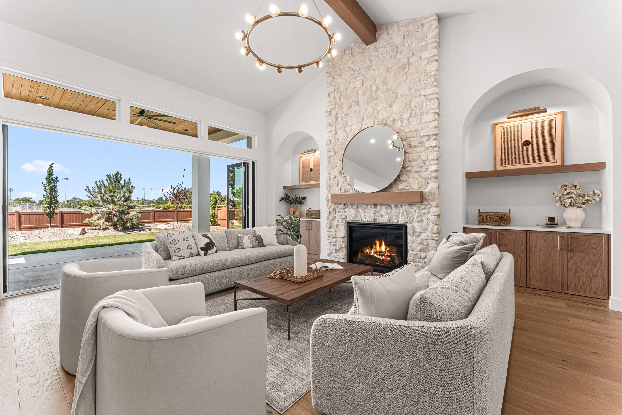 Interior of a modern living room with white walls, large sliding glass doors, a stone fireplace, circular and rectangular mirrors, and cozy seating including white and gray chairs and sofas with decorative pillows. There's a wooden coffee table with a candle and a tray, and built-in shelves with artwork, decor, and a vase with flowers. The room is bright and welcoming, with a view of the backyard and outdoor covered patio.