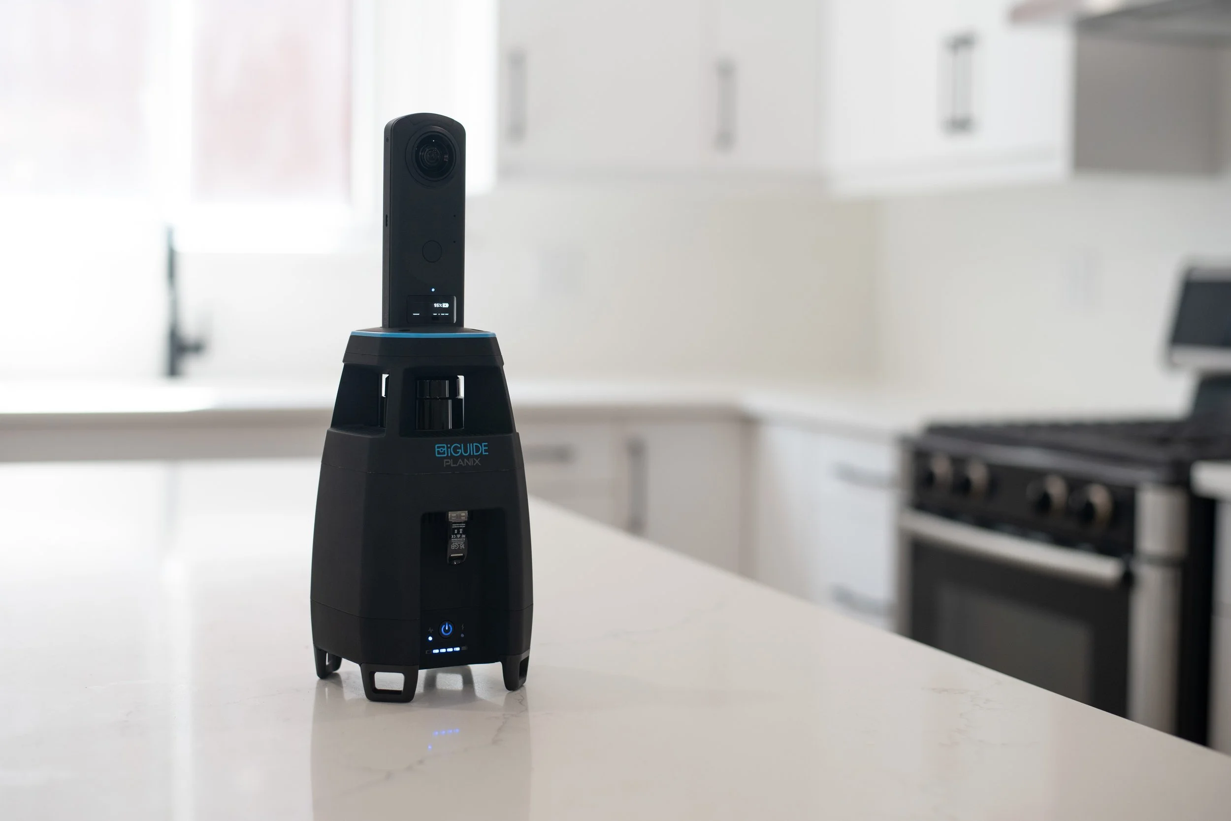 A black 3D scanner device on a white countertop in a bright, modern kitchen with white cabinets and a stove in the background.