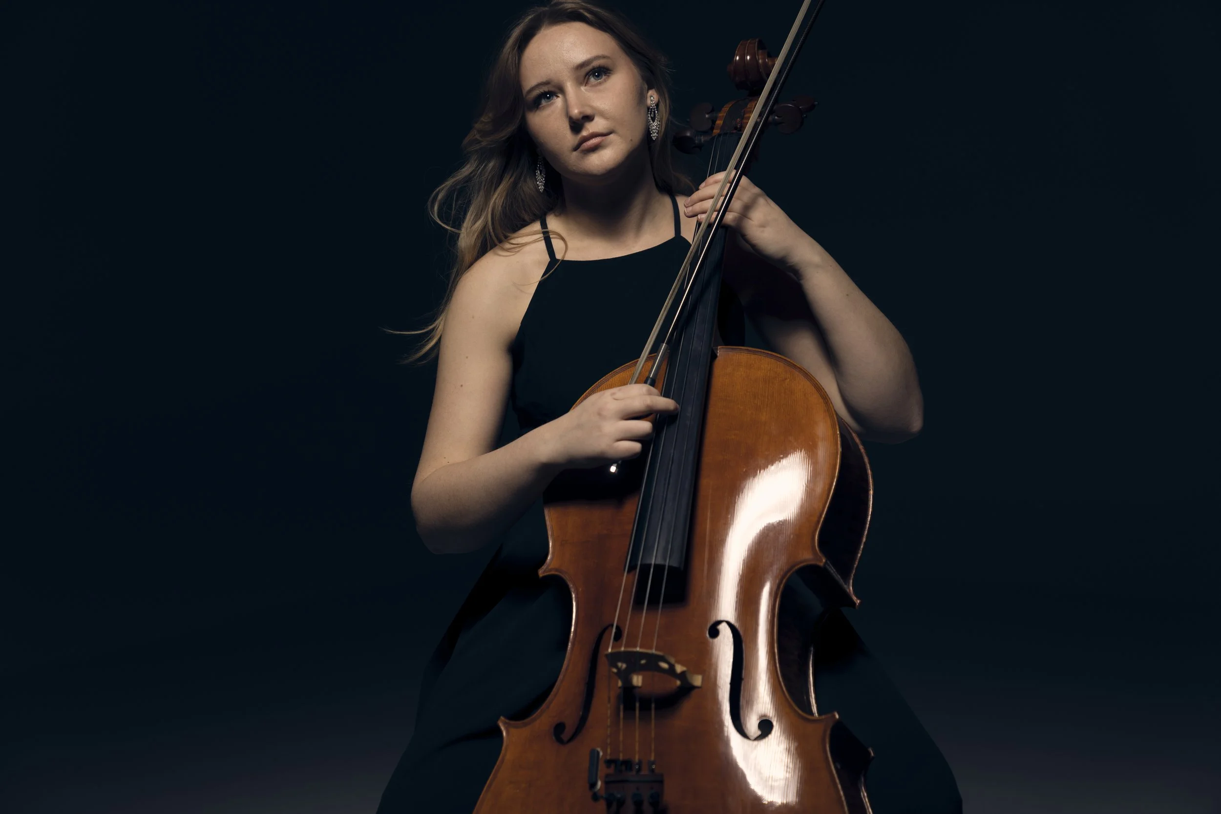 A woman with brown hair is playing a wooden cello against a dark background.