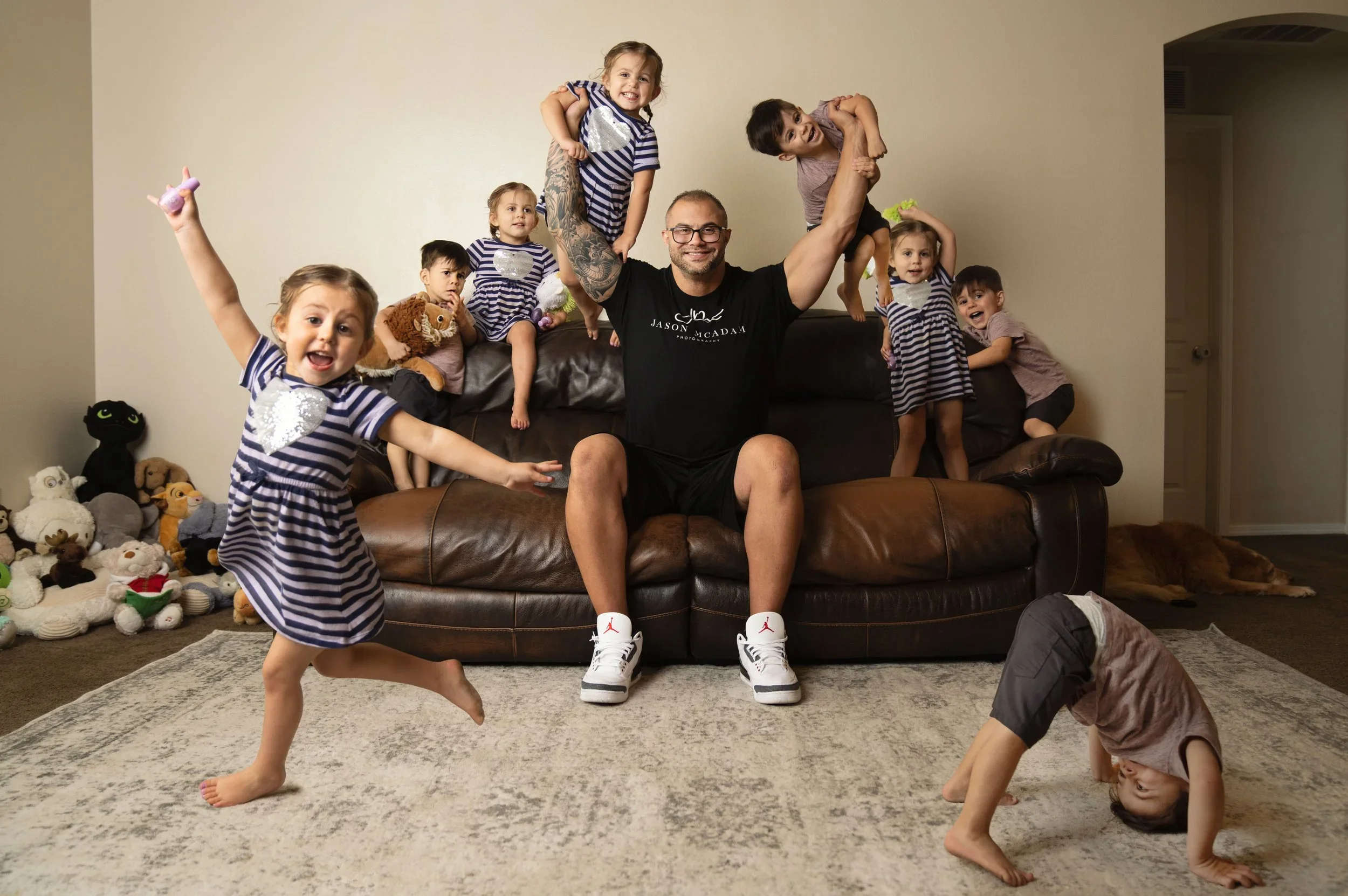A man with glasses and a beard, sitting on a leather couch, holding two young children on his arms. Several children are engaging in various activities around him, with some posing for the camera and others playing on the floor. There are stuffed animals on the floor and a plain wall in the background.