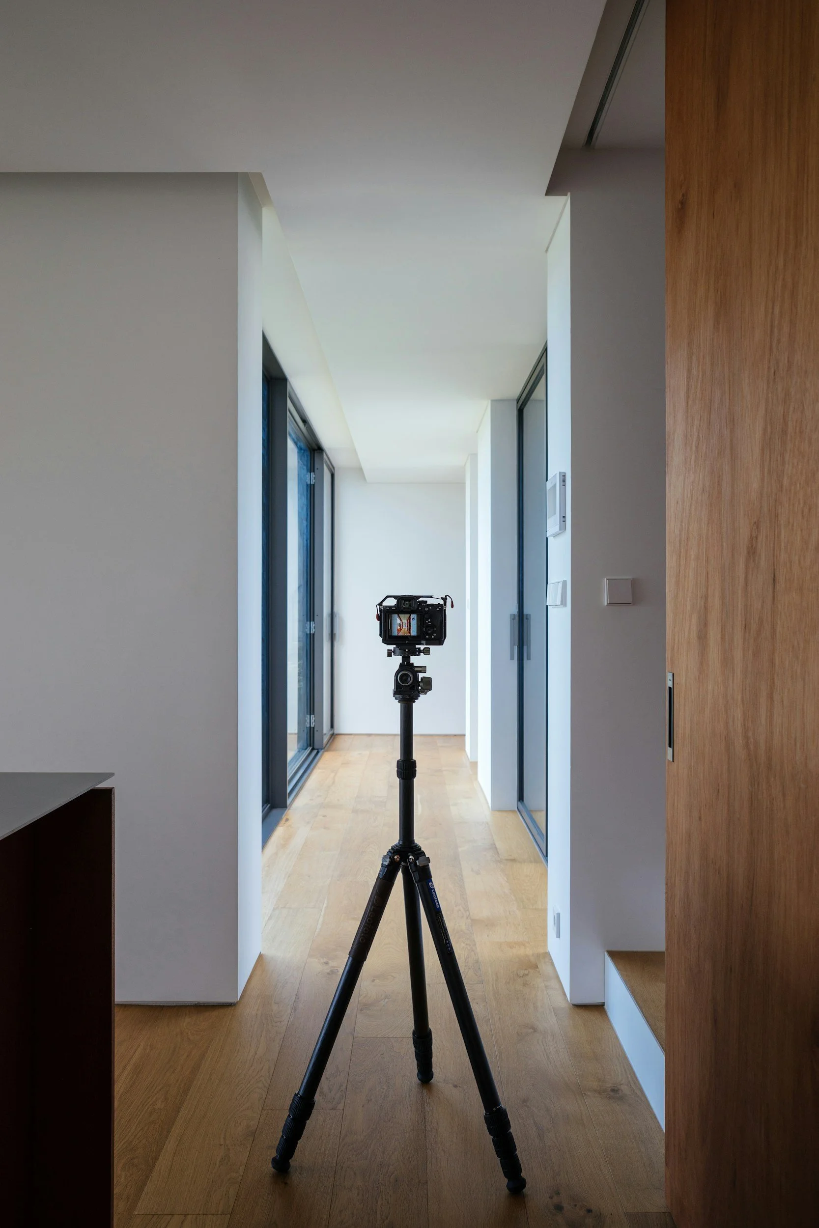 Photo of a camera on a tripod set up inside a modern apartment or house, with large windows along the hallway and wooden flooring, seen from the entrance.