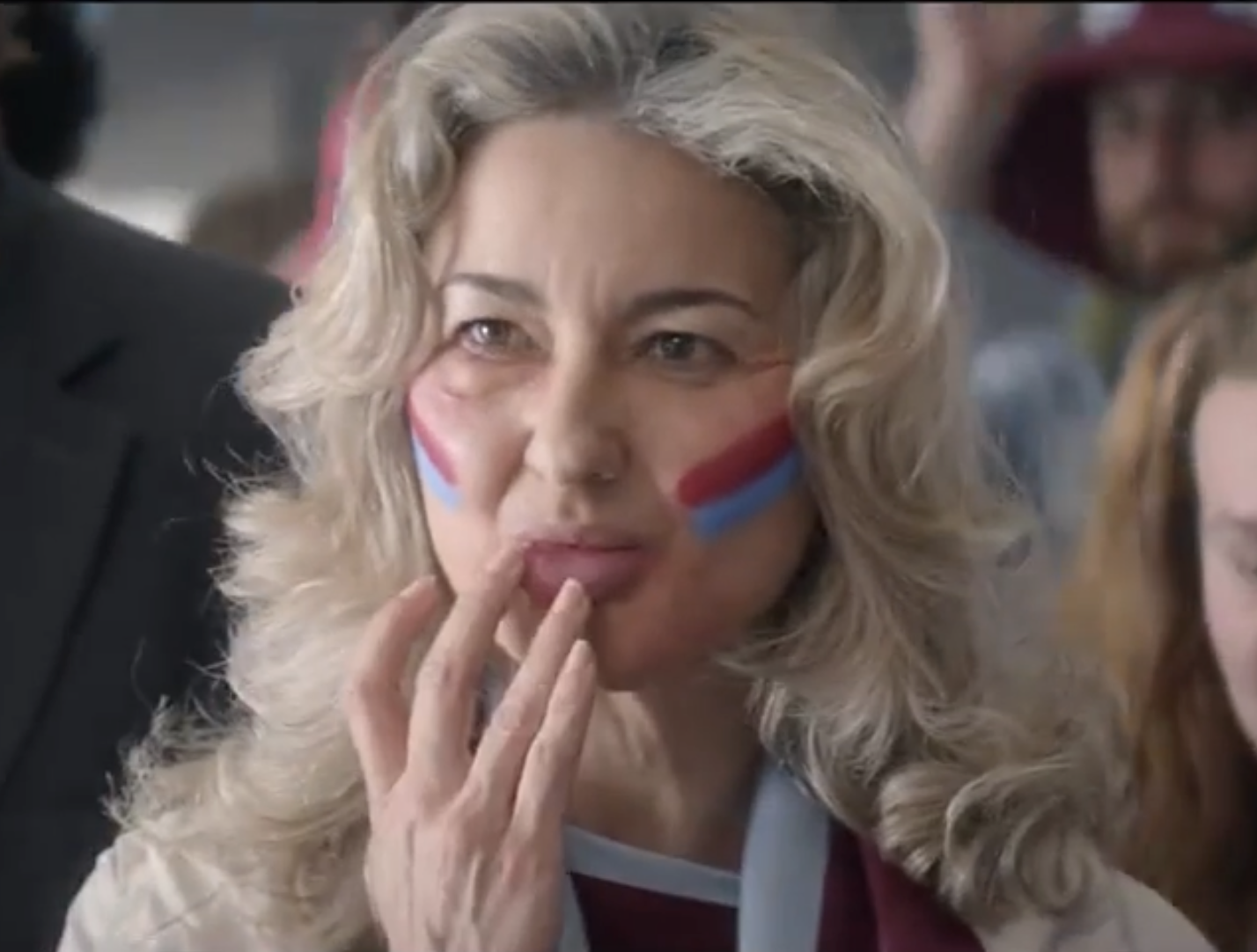 Woman with blonde hair and face paint in red and blue, looking contemplative with fingers on lips at a crowded event.