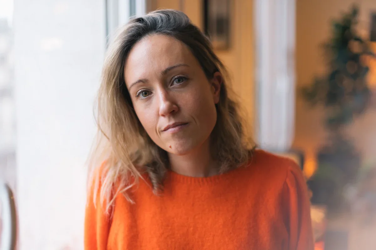 A woman with shoulder-length blonde hair wearing an orange sweater looks at the camera indoors near a window with natural light.
