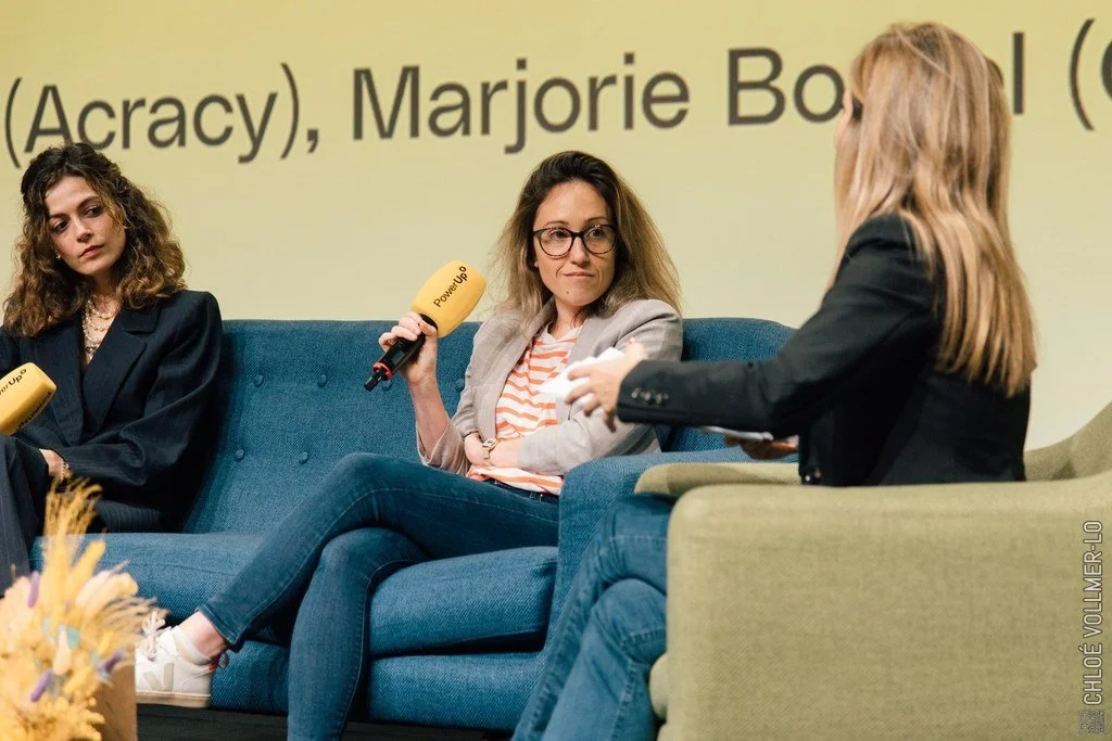 Three women sitting on a blue and green couch during a panel discussion, with a yellow background displaying partially visible text.