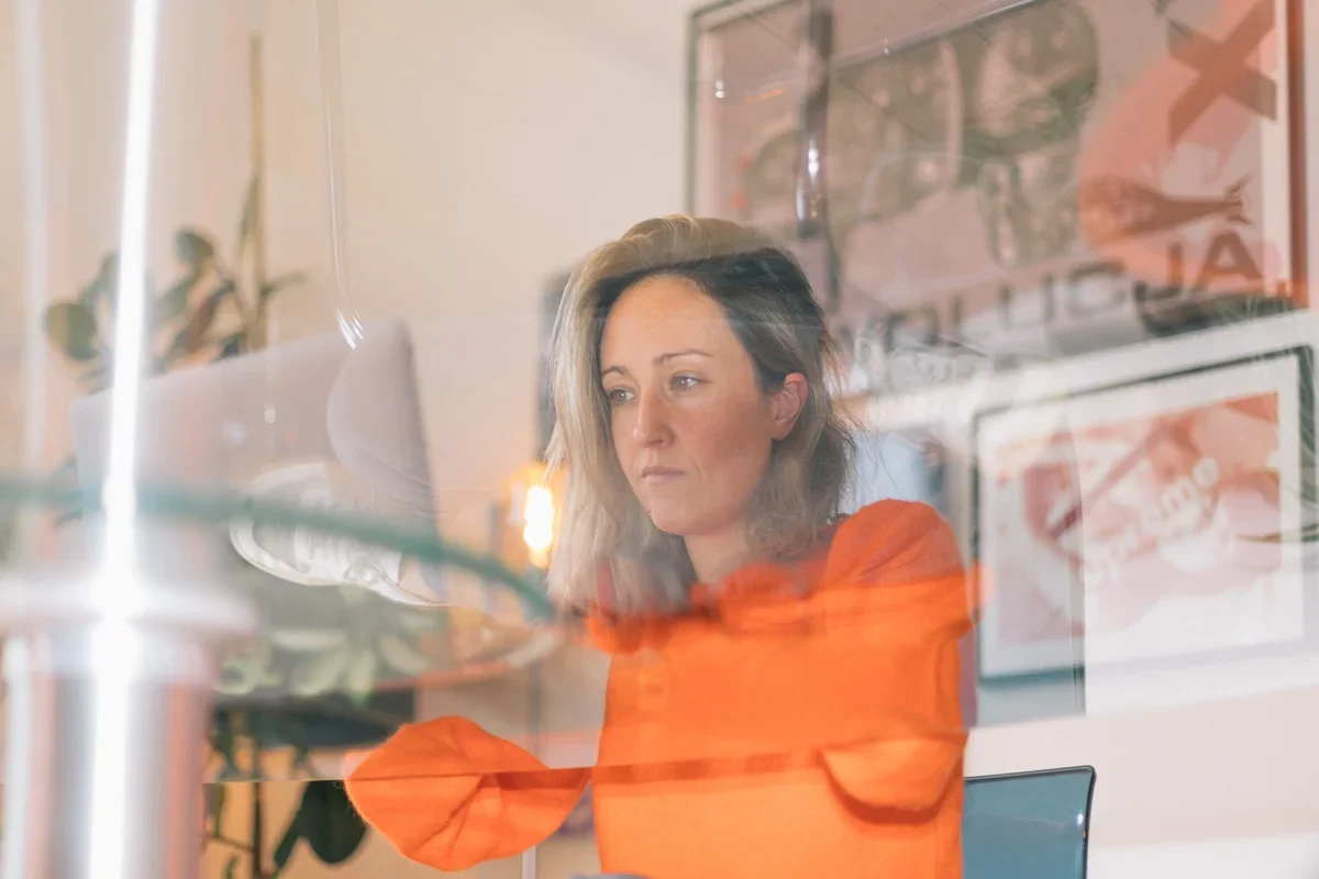 A woman with blonde hair wearing an orange top looking at a computer screen through a glass partition in an office setting.