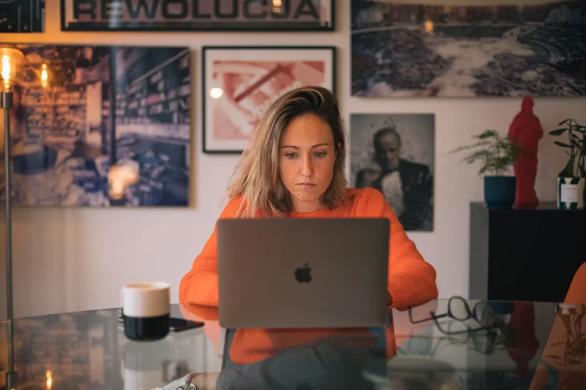 A woman working at a desk with a laptop, glasses, a coffee mug, and artwork on the wall behind her in a cozy room.
