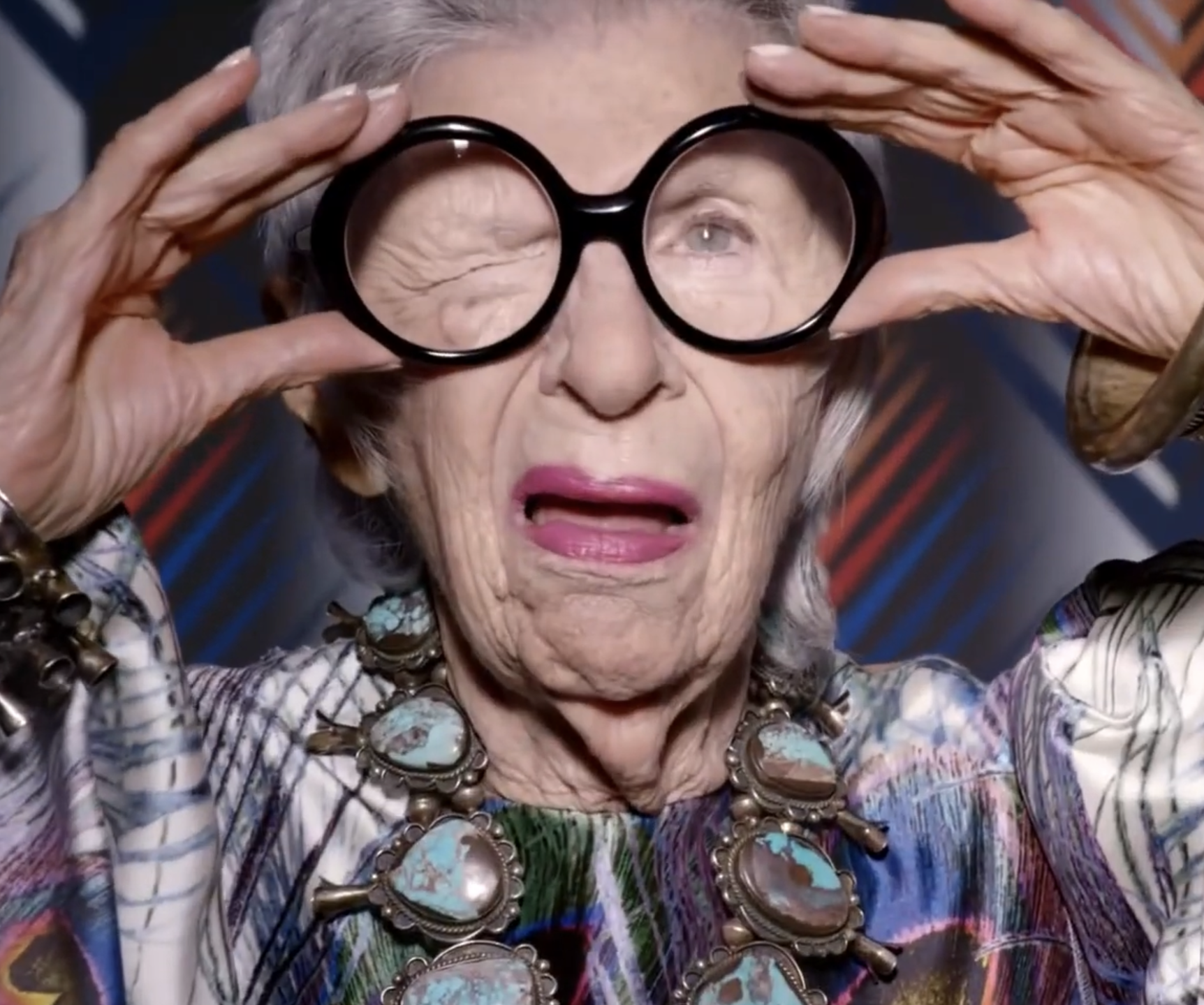 Elderly woman with gray hair and bright pink lipstick, wearing a colorful top and large turquoise jewelry, holds black eyeglasses in front of her eyes with an expressive facial expression.
