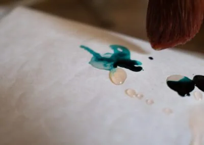 Close-up of a person's finger applying blue and black watercolor or ink paint onto white paper.