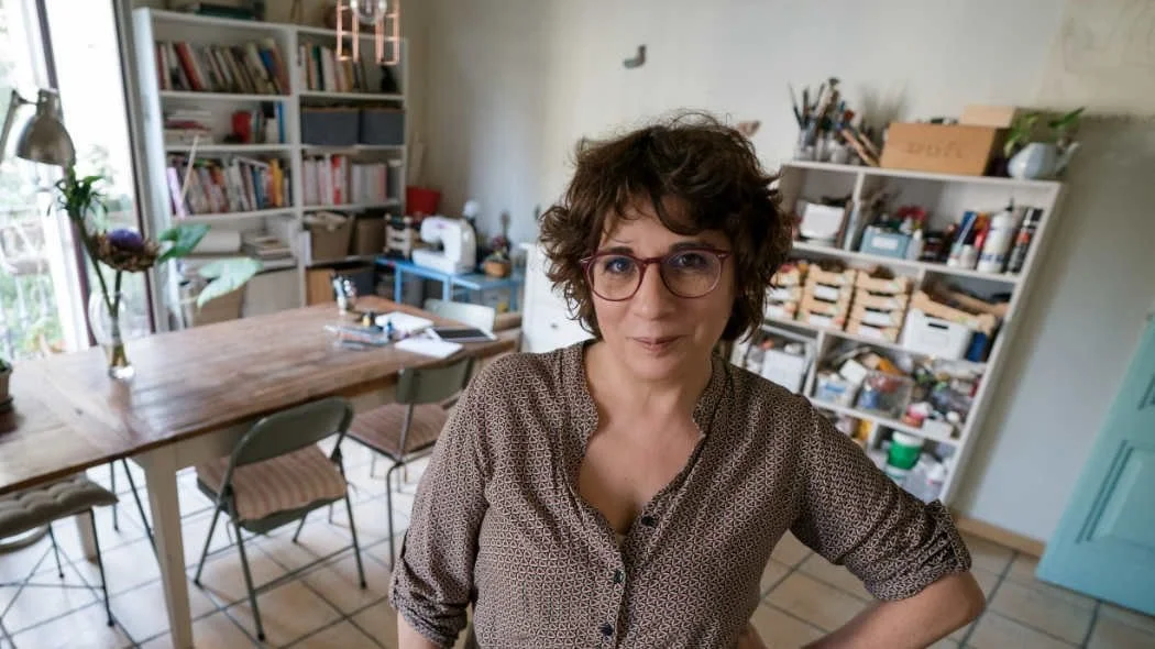 A woman with curly brown hair and glasses standing in a well-lit kitchen or craft room, with bookshelves and a wooden dining table in the background.