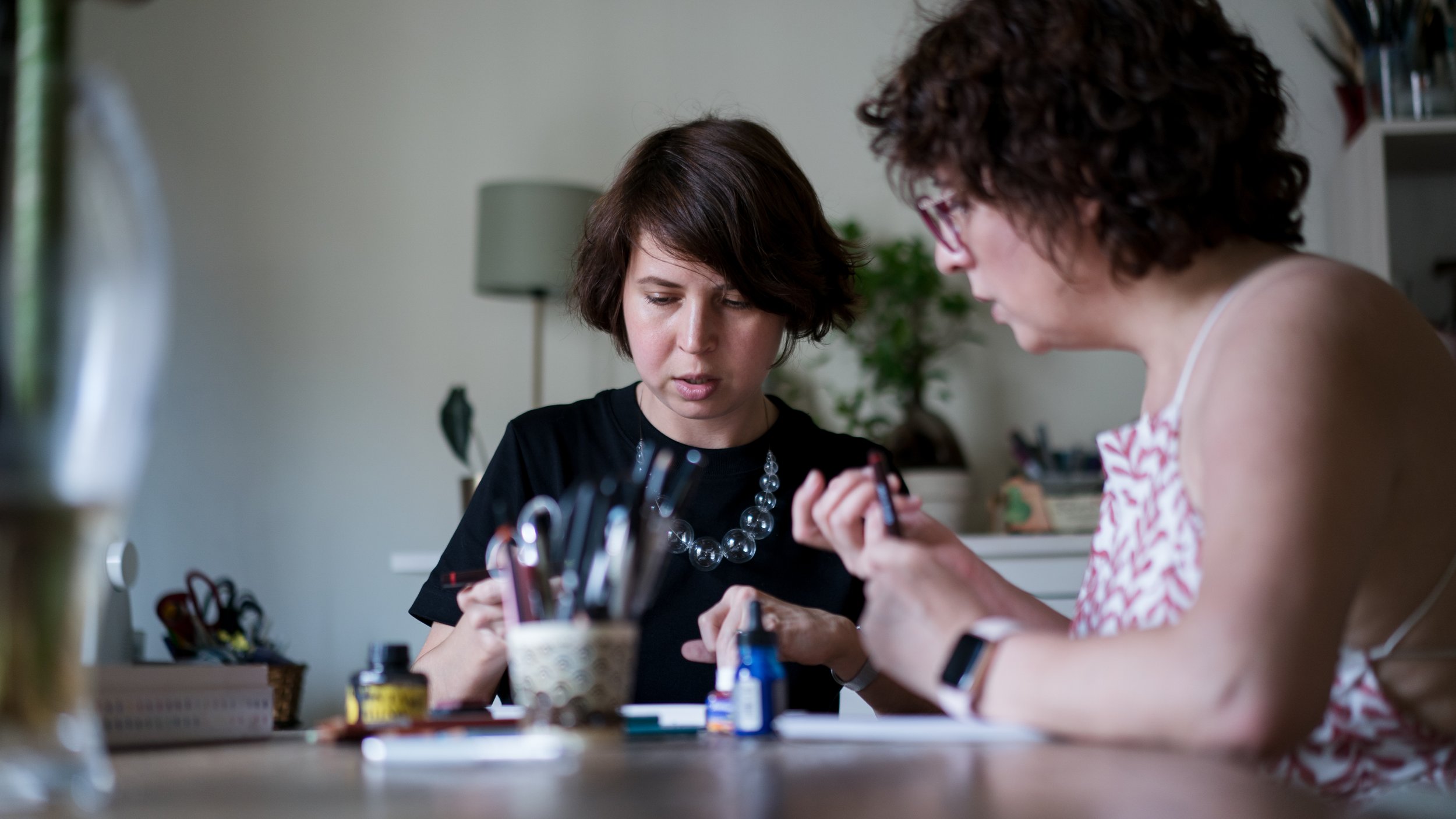 Two women with short brown curly hair examining nail polish at a table, with nail tools and bottles in front of them, in a room with a lamp and plants in the background.