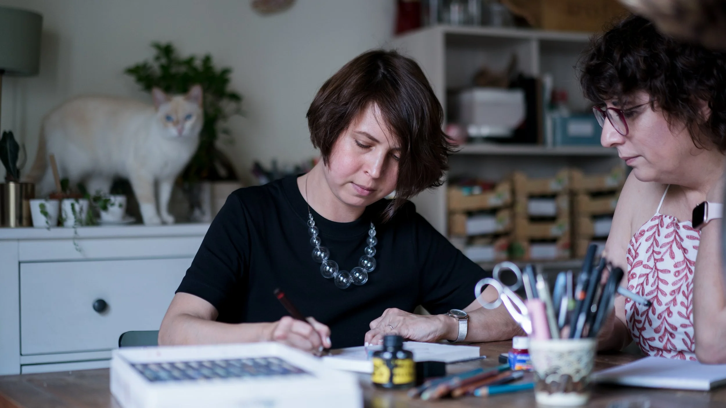 Two women sitting at a table engaged in artistic work, with art supplies like paints, brushes, and palettes in front of them. A cat stands on a white cabinet in the background.