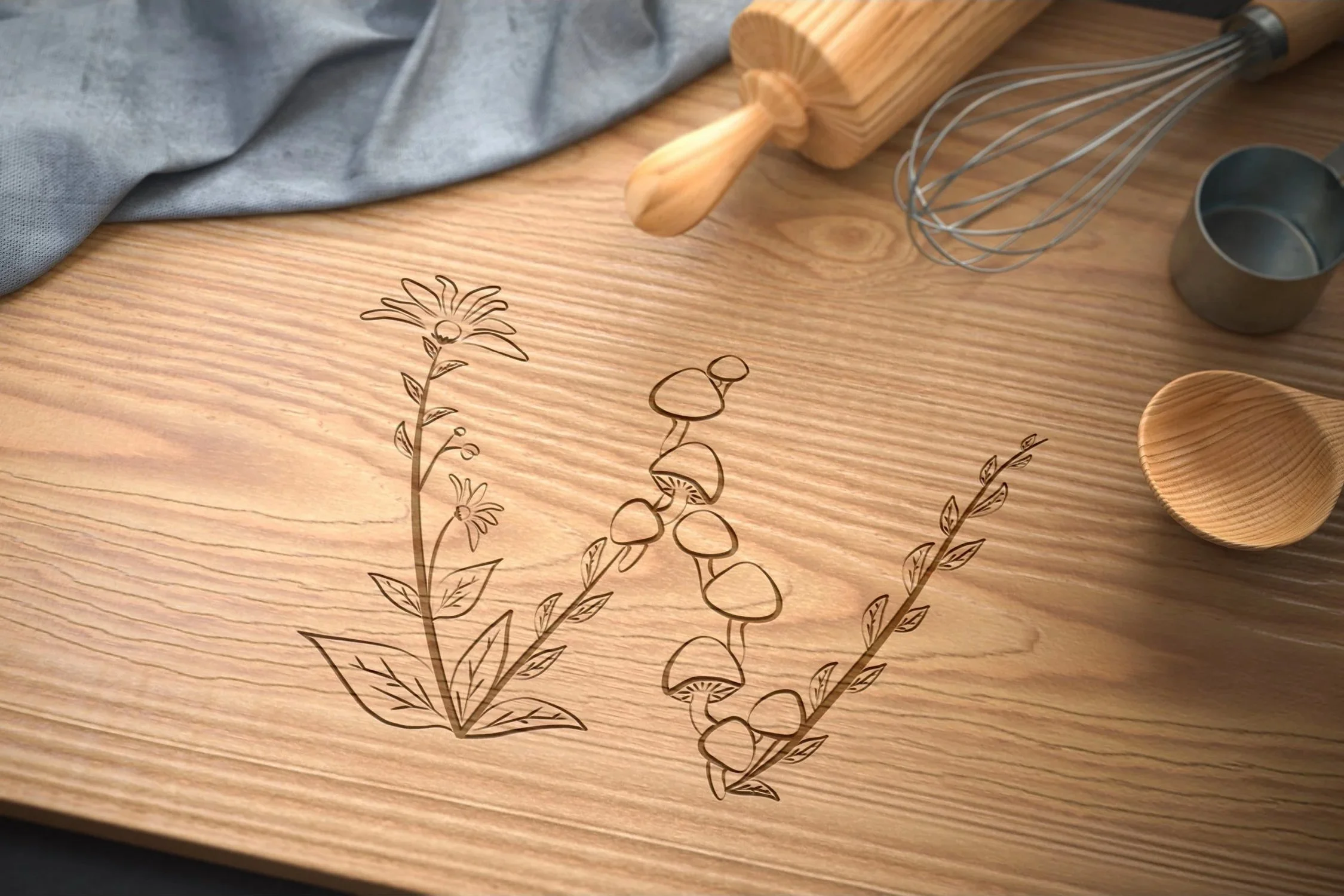 A wooden cutting board decorated with black line art illustrations of mushrooms and plants, with a gray cloth, a wooden rolling pin, a metal whisk, and a metal measuring cup placed on it.