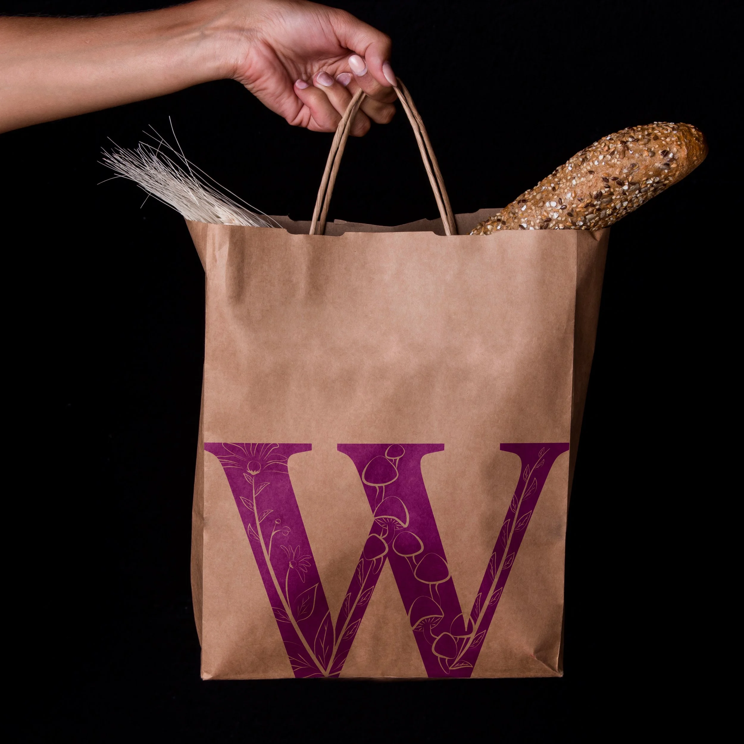 A hand holding a paper bag with a loaf of multigrain bread sticking out of it, against a black background.