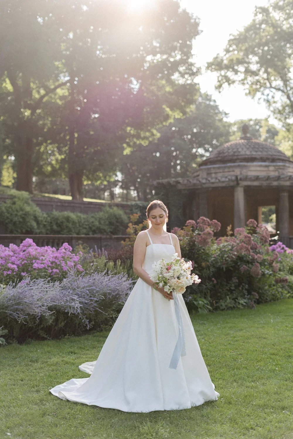 A bride in a white wedding dress holding a bouquet of white and pink flowers, standing on a well-manicured lawn with pink and purple flowering shrubs, surrounded by trees, with a gazebo in the background and sunlight filtering through the trees.