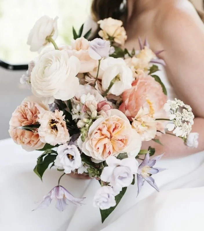 Close-up of a bouquet of pastel-colored flowers including peonies, roses, and other blooms, with a person holding it with part of their arm visible.
