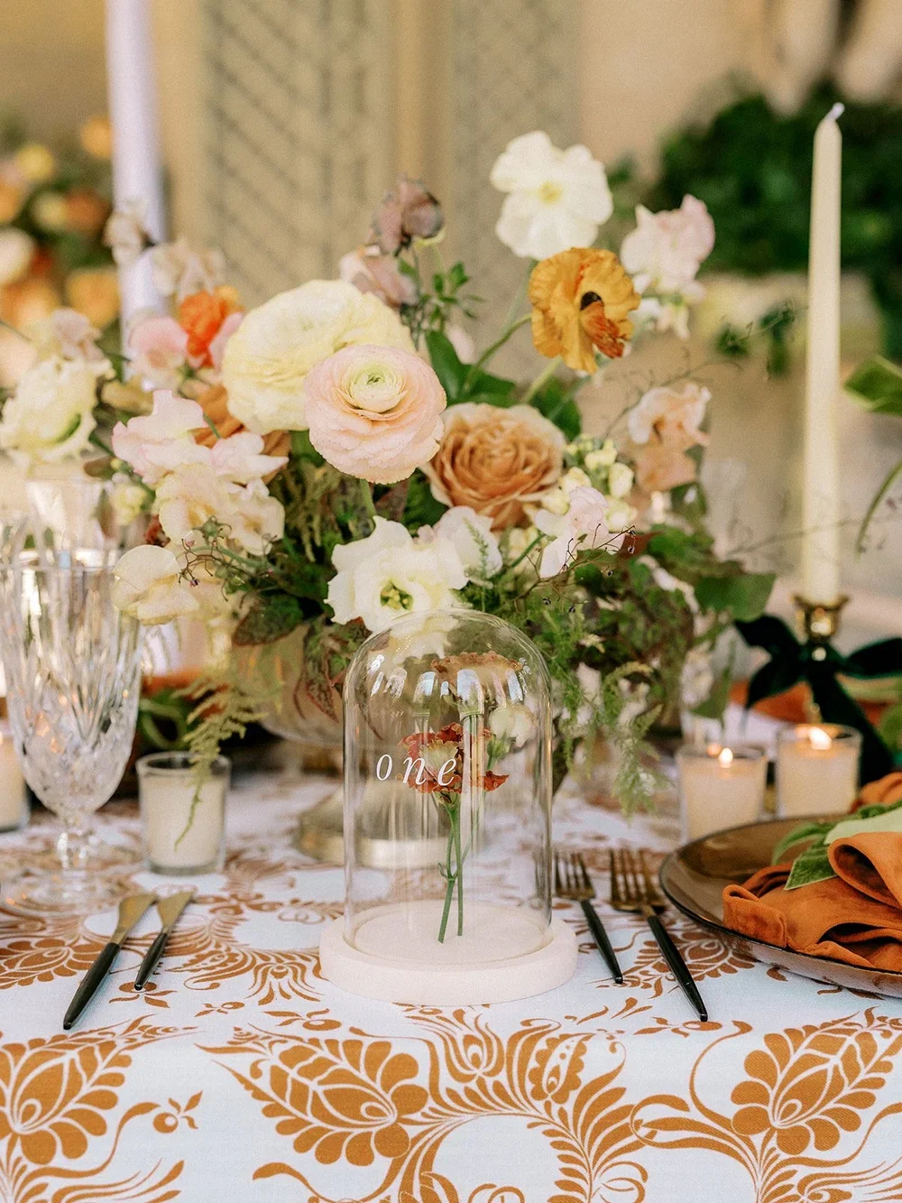 A decorated table with a floral centerpiece, candles, glasses, and cutlery, with a glass dome with "one" written on it over a flower inside.