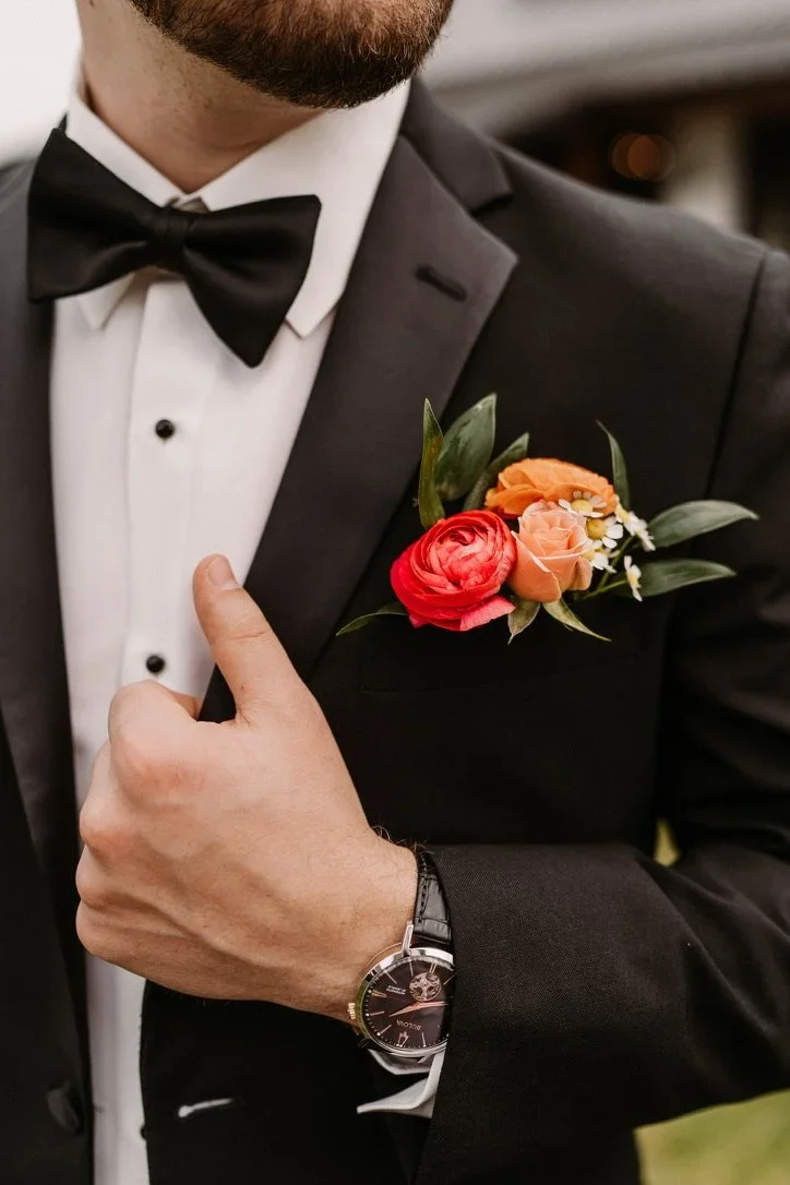 Close-up of a man in a tuxedo with a black bow tie, adjusting his jacket to reveal a colorful boutonnière with red, orange, and white flowers, wearing a black leather strap watch.