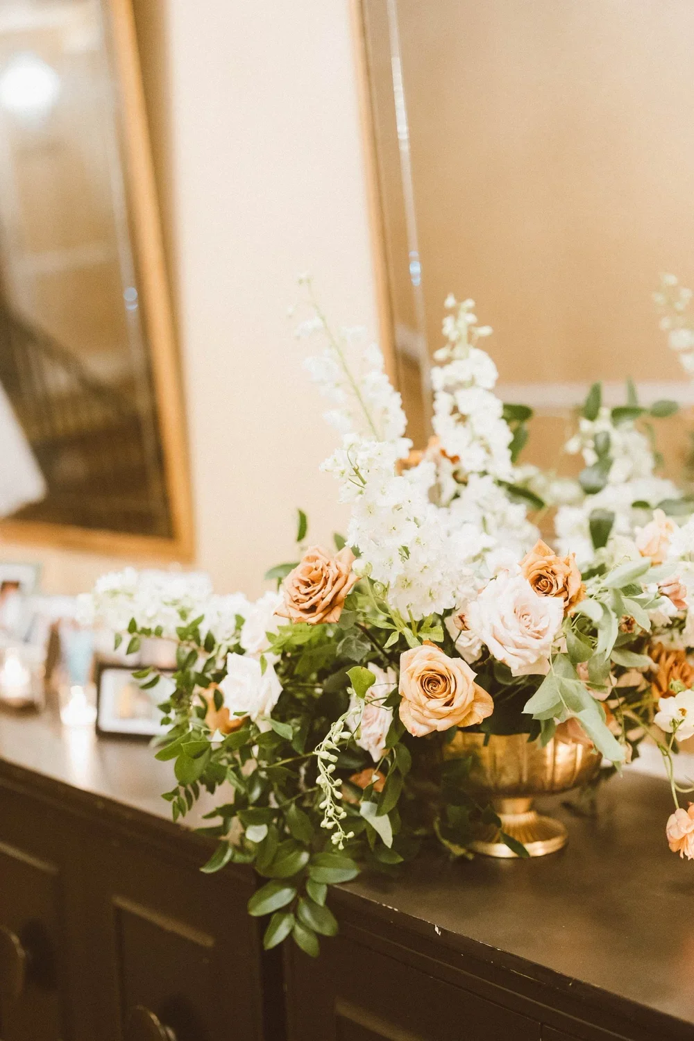 A floral arrangement with peach and white roses, white snapdragons, and green foliage in a gold bowl on a dark wooden surface, with framed photos and mirrors in the background.