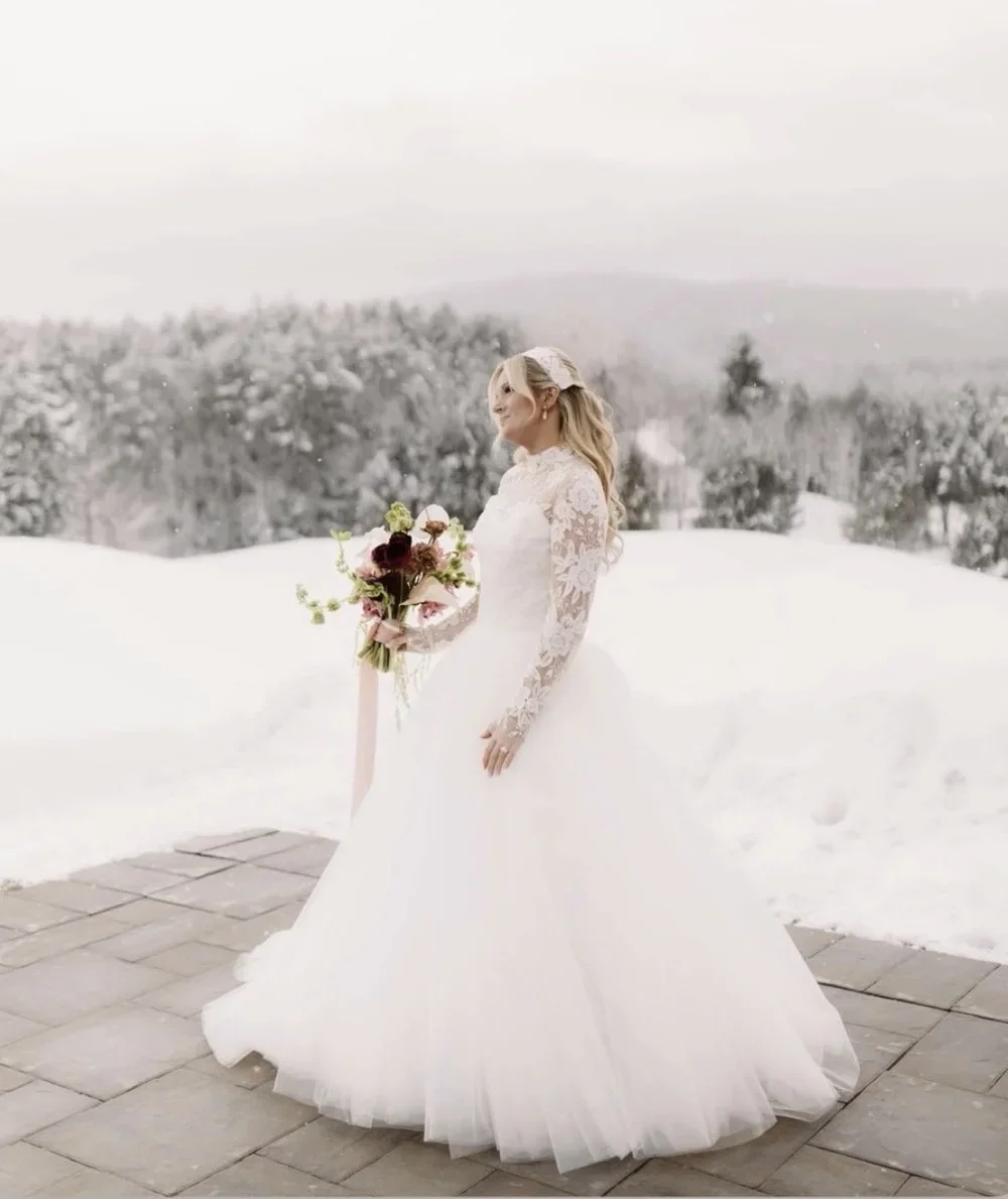 A bride in a white wedding dress with lace sleeves holding a bouquet of pink, white, and red flowers in a snowy outdoor landscape.