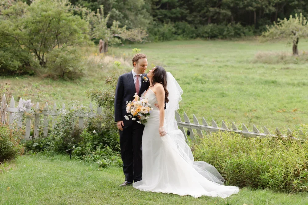 A bride and groom stand close together in a lush green garden, with the bride holding a bouquet of flowers and smiling at each other.