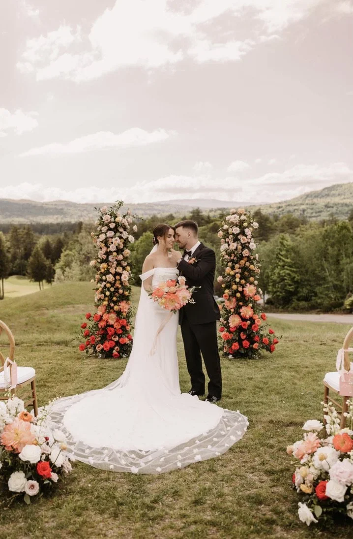 Bride and groom standing close together during outdoor wedding ceremony with floral arch behind them and floral arrangements on the ground, green rolling hills in the background.
