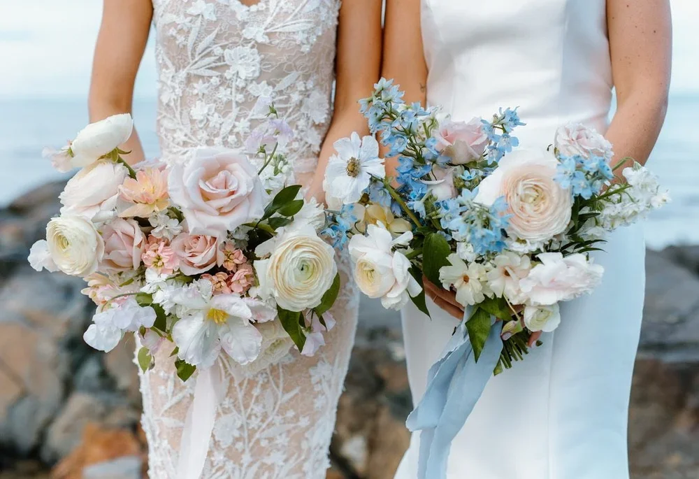 Two women in wedding dresses holding bouquets of pink, white, and blue flowers outdoors near the coast.