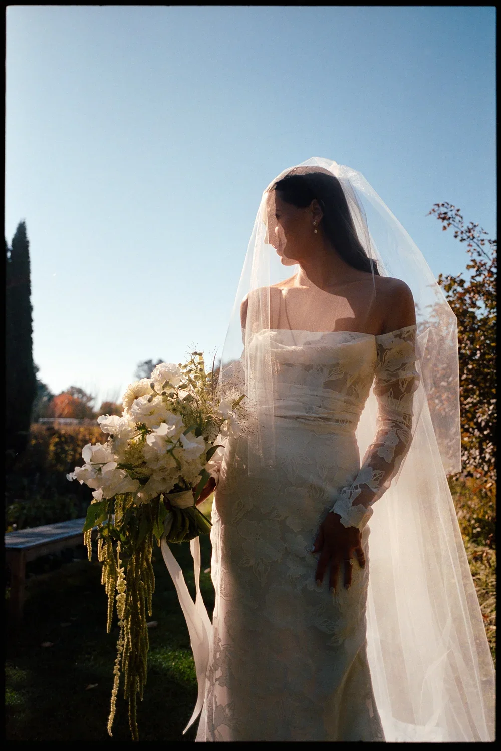 Bride in an off-the-shoulder lace wedding dress and veil holding a bouquet of white flowers, standing outdoors during sunset.