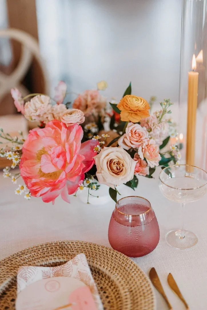 A table centerpiece with a bouquet of pink, cream, and peach flowers, a pink glass candle holder, and two empty glasses. The table is set with a woven placemat, a napkin, and gold utensils. There is a lit candle in the background.