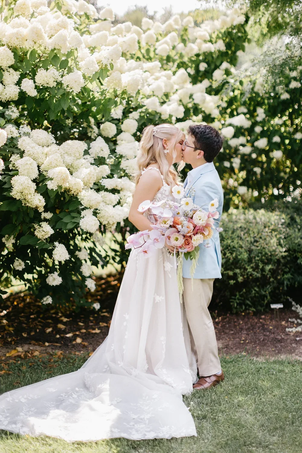 A bride and groom sharing a kiss outdoors in front of a large white flowering bush, with the bride holding a bouquet of pink and white flowers.