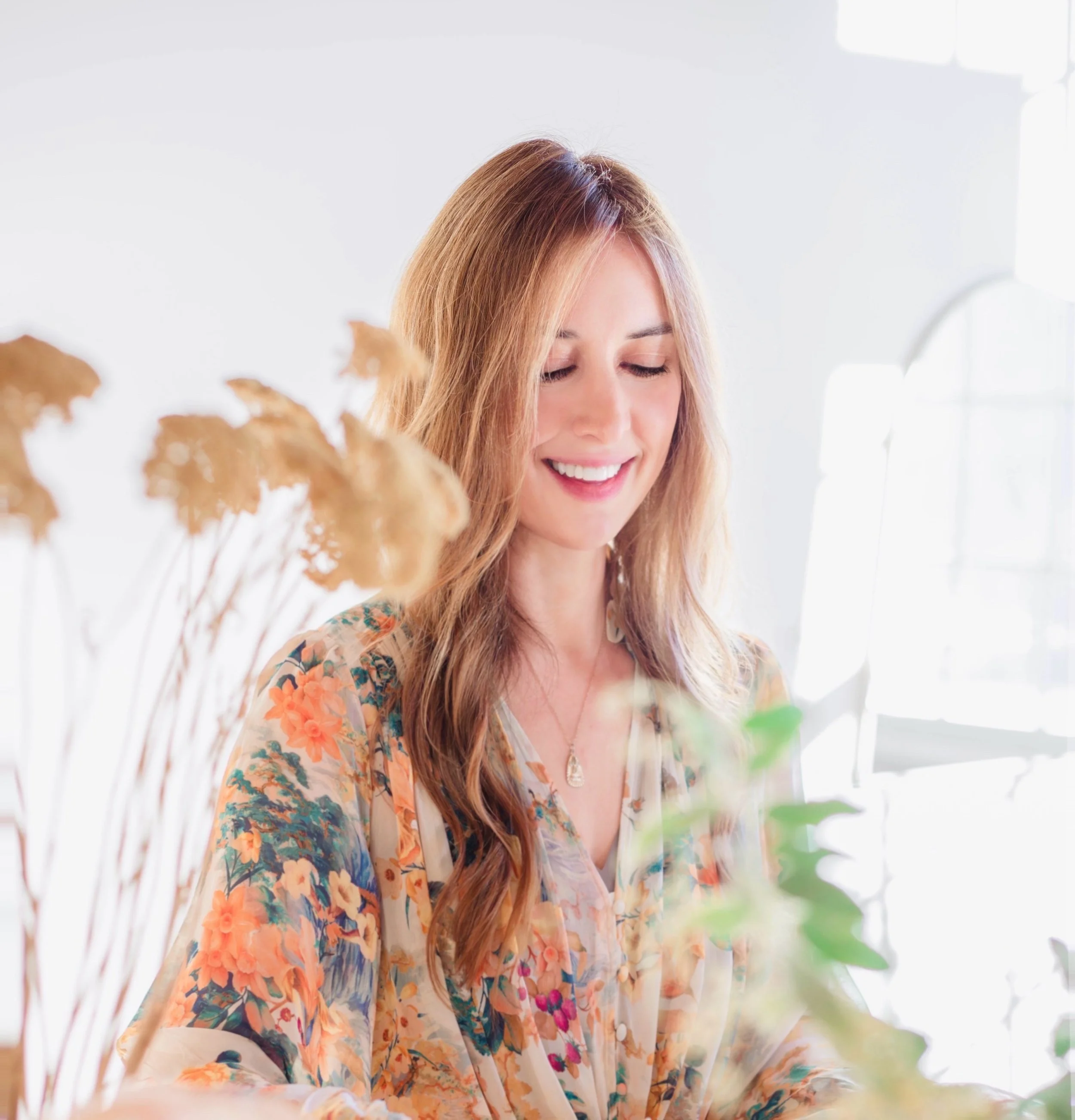 A woman with long wavy hair smiling with closed eyes, wearing a floral blouse, in a bright room with natural light.