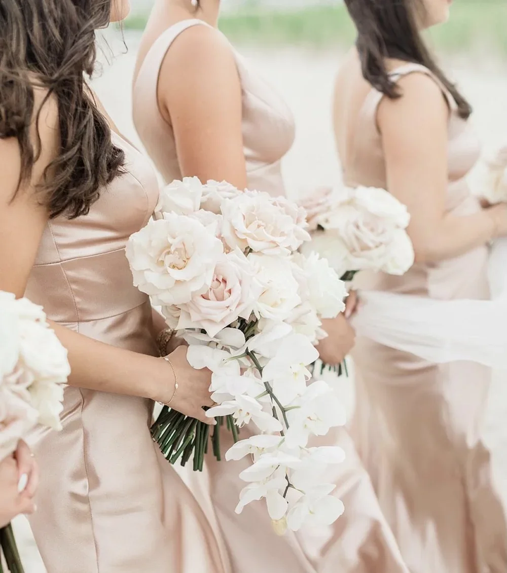 Three women in beige satin dresses holding bouquets of white and blush pink flowers, possibly at a wedding or formal event.