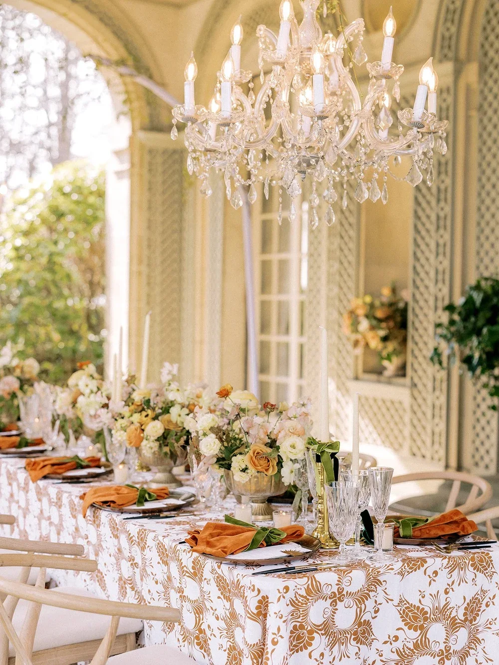 Elegant outdoor dining table set under a chandelier with floral arrangements and candles.