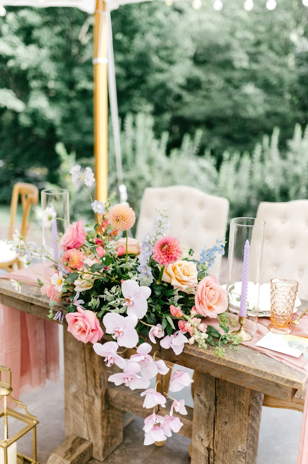 Wedding table centerpiece with pink and peach roses, white orchids, and light purple flowers on a rustic wooden table outdoors.