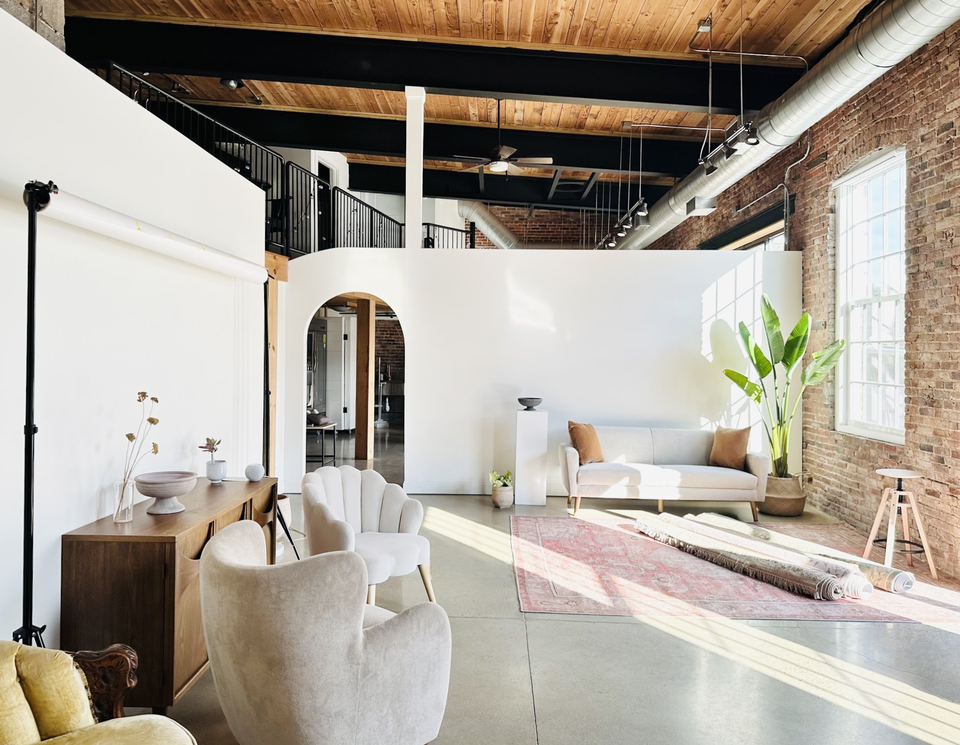Bright industrial-style living room with exposed brick walls, large windows, a white sofa, a vintage armchair, a wooden sideboard with decor, a pink area rug, and a large potted plant.