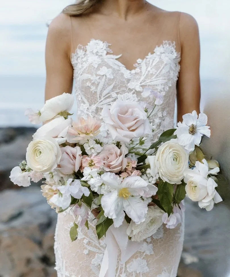 A woman in a lace wedding dress holding a large bouquet of white and pale pink flowers outdoors.