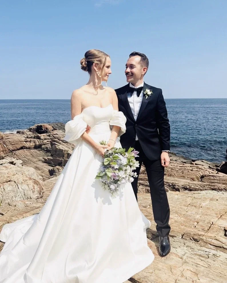 Bride and groom standing on rocks by the ocean during their wedding, smiling at each other with the bride holding a bouquet of flowers.