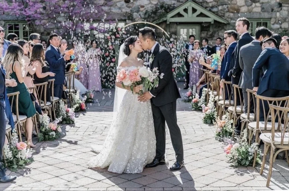 Bride and groom kissing at their wedding ceremony with guests clapping and celebrating around them, outdoor setting decorated with flowers.