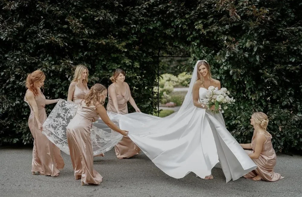 A bride with a bouquet standing on her wedding gown while five bridesmaids help her with the dress outside, with greenery in the background.