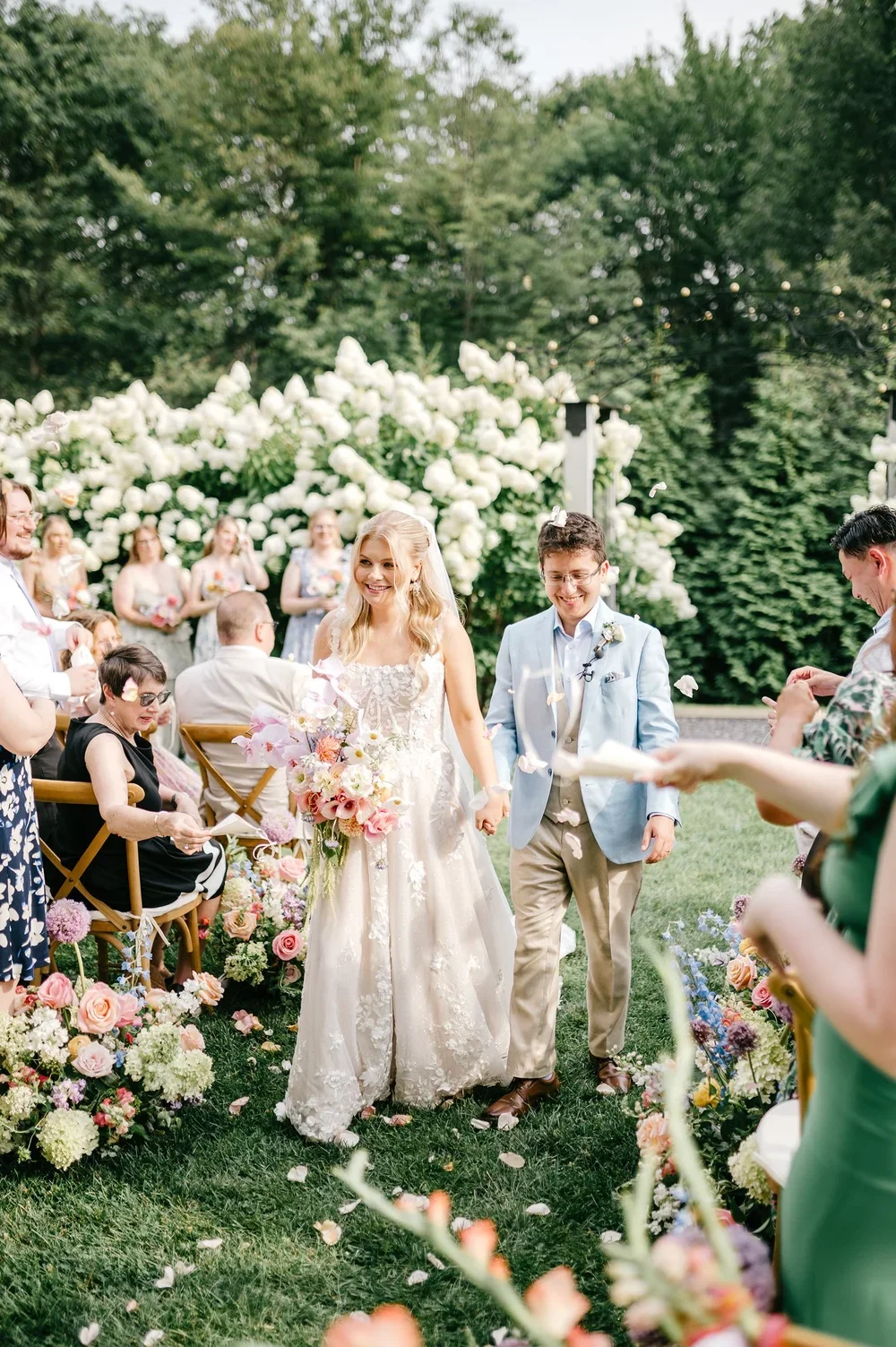 Bride and groom walking hand-in-hand down the aisle at an outdoor wedding, surrounded by friends and family, with a floral arch and greenery in the background.