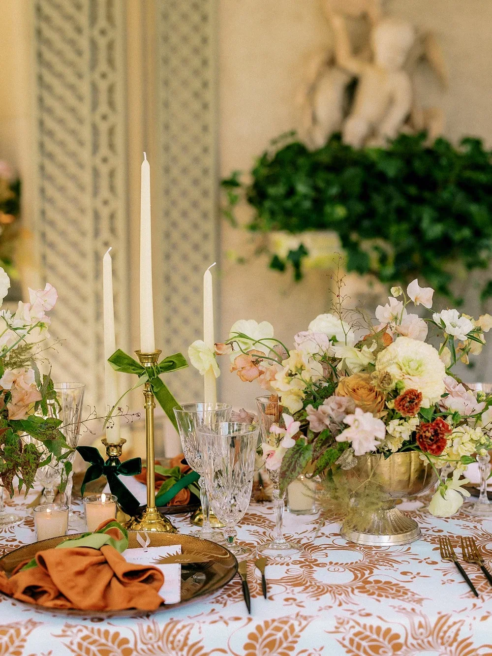 Elegant dining table setup with floral centerpiece, gold candlesticks with white candles, crystal glassware, gold-rimmed plates, and orange cloth napkins, in a decorated indoor setting.