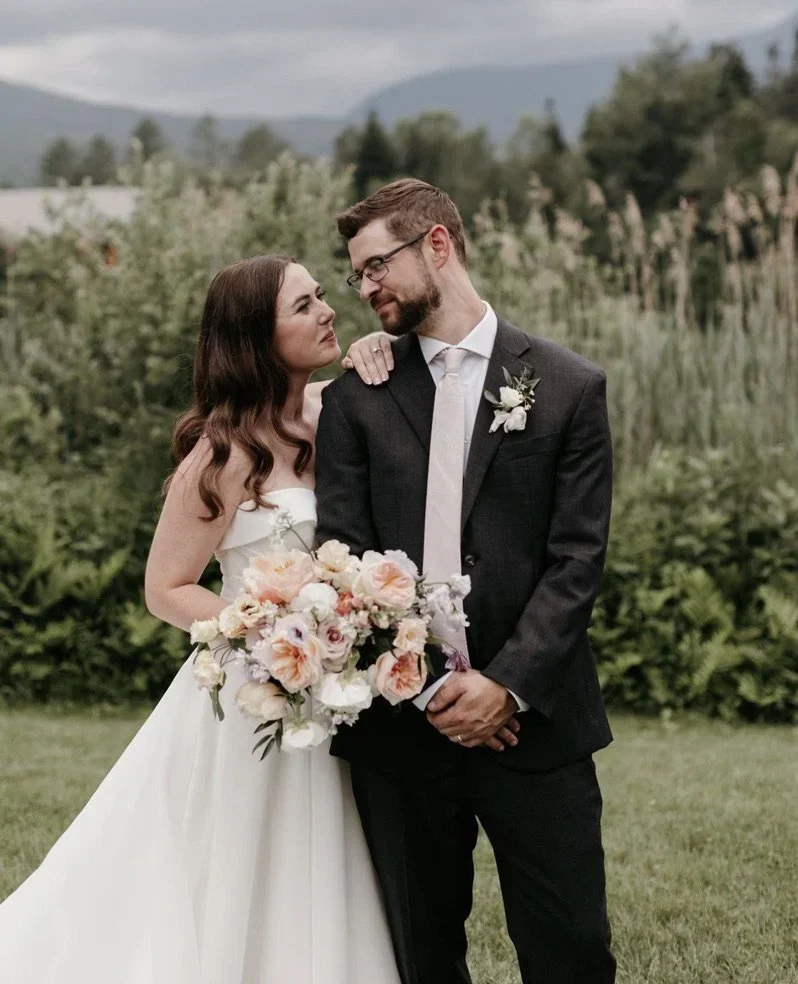 A bride and groom standing outdoors, gazing at each other, with the bride holding a large bouquet of flowers, and suited in wedding attire.