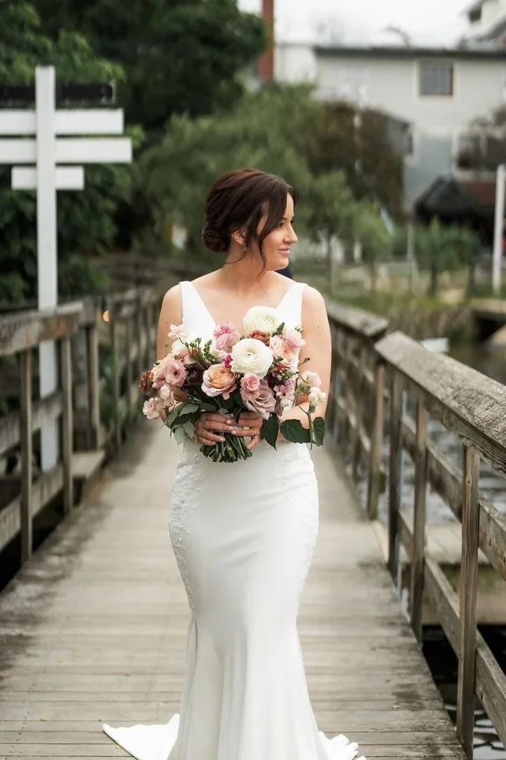A bride in a white wedding dress holding a bouquet of pink, white, and peach flowers walking on a wooden bridge outdoors.