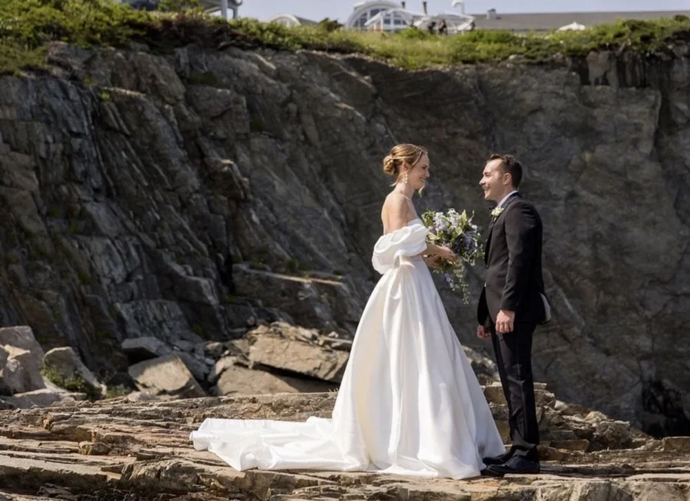 A bride and groom standing on rocks during their wedding photoshoot, with a rocky cliff in the background and some cars visible at the top of the cliff.
