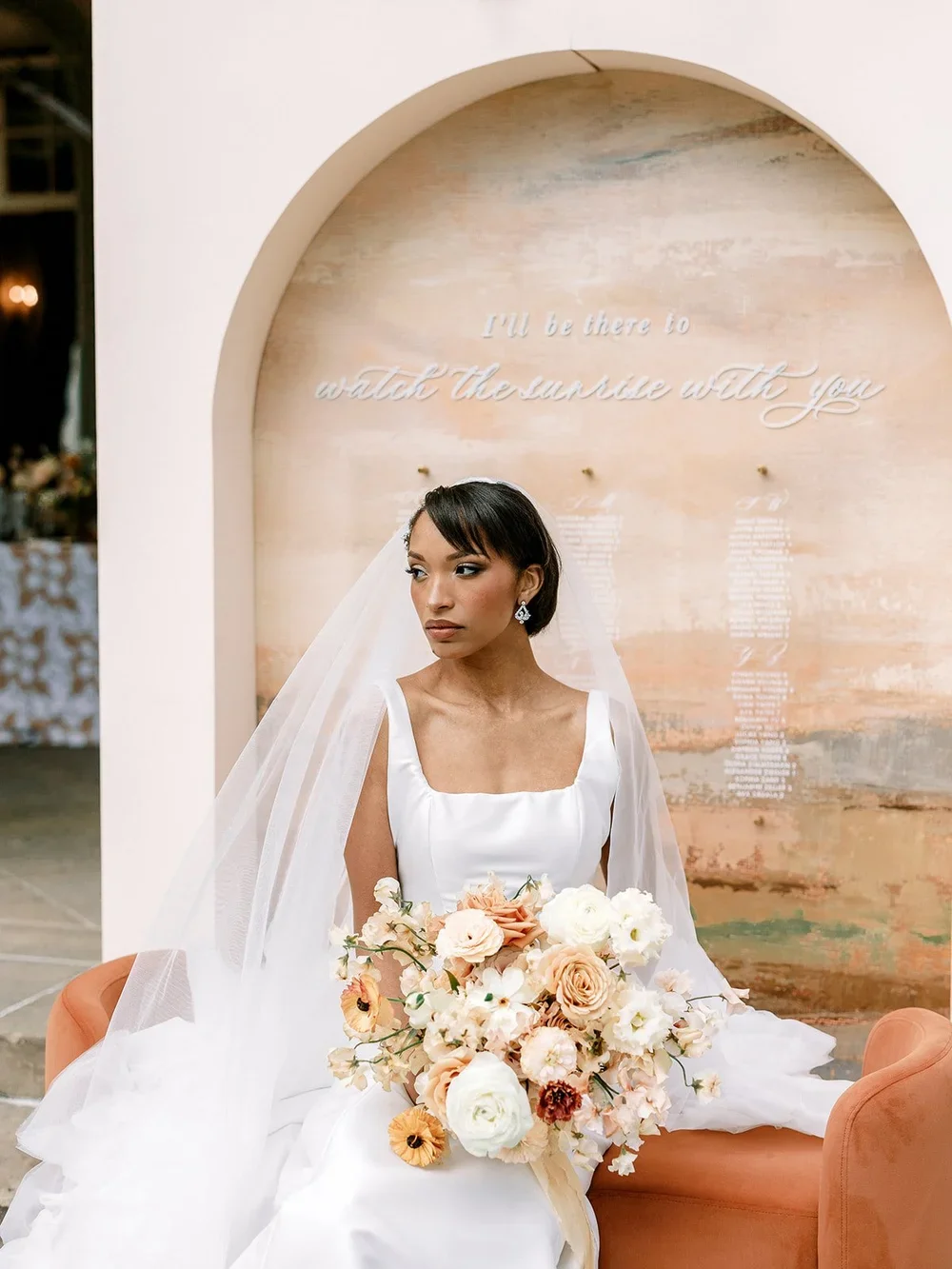 A bride in a white wedding dress sitting on a peach-colored chair, holding a large bouquet of light-colored flowers. She is wearing earrings and a veil, with a contemplative expression, in front of a textured wall with cursive text.
