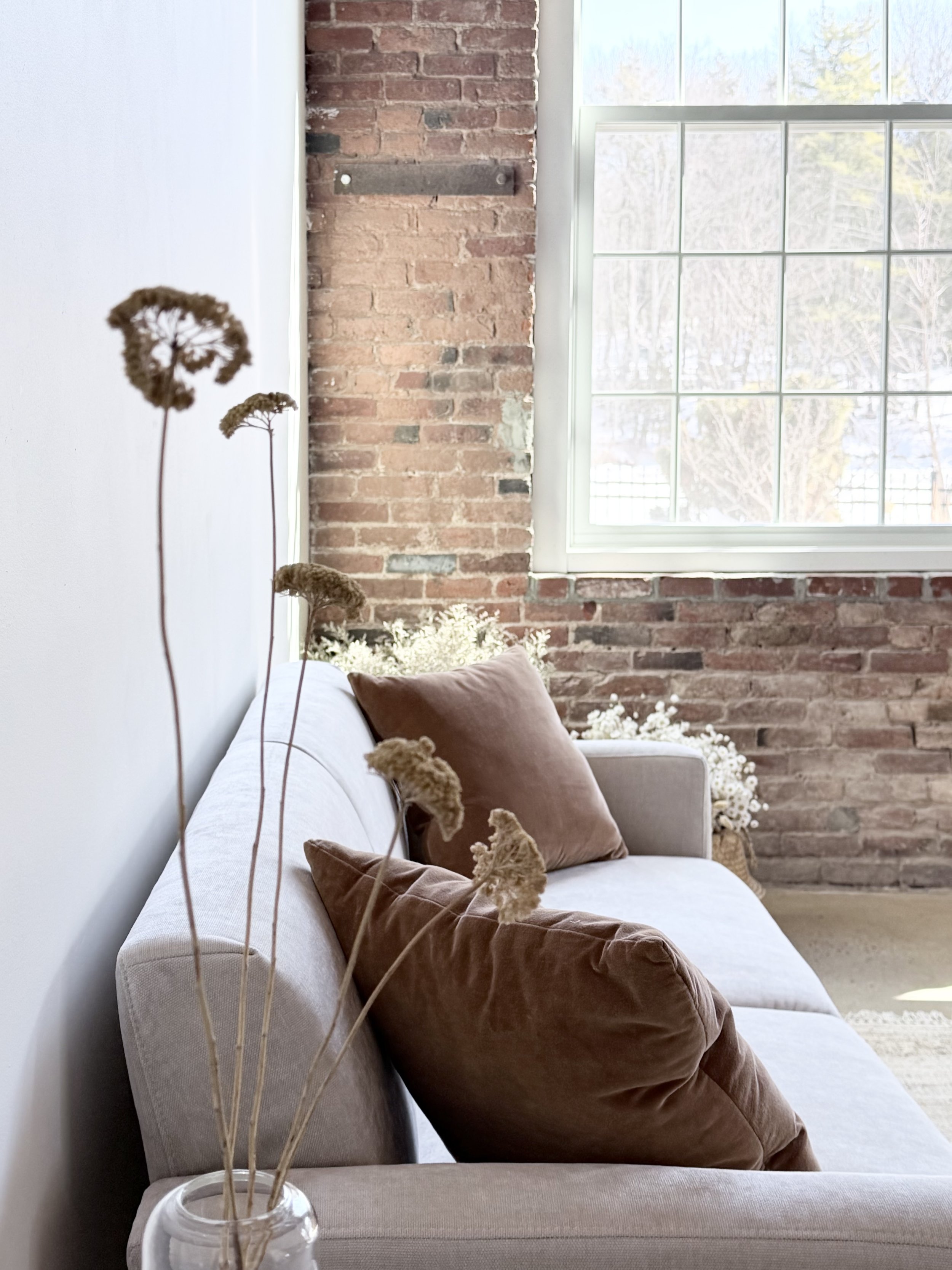 Living room with a beige sofa, brown pillows, dried flowers in a white vase, and a brick wall by a large window showing an outdoor view.