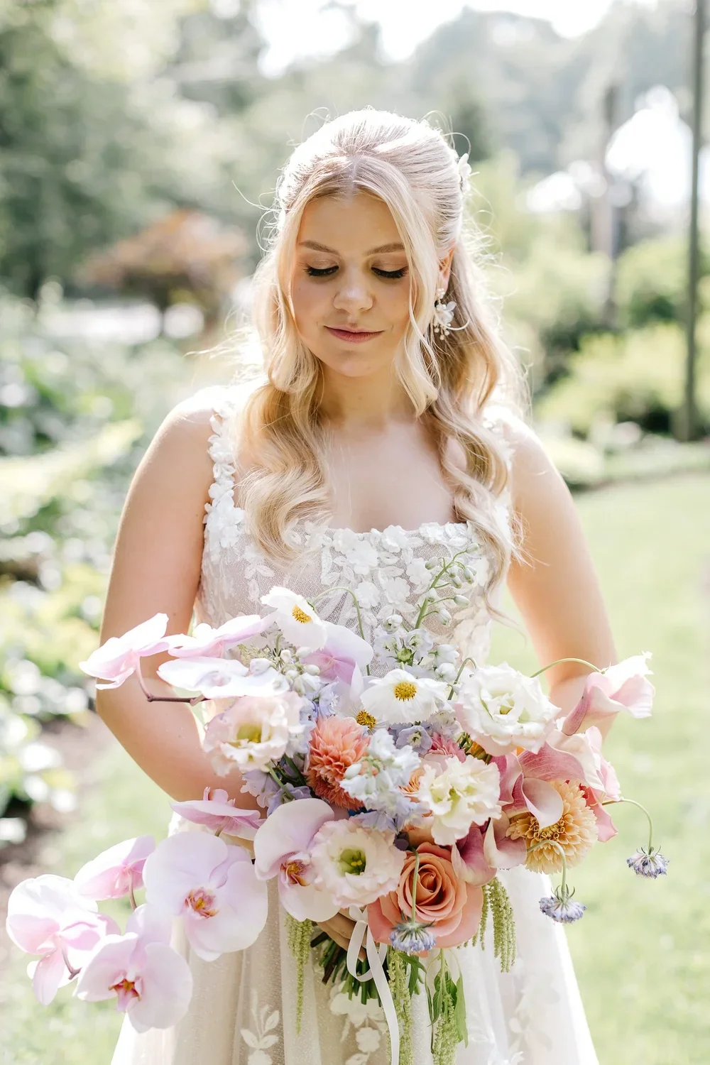 A bride with long blonde hair in loose curls, wearing a white lace wedding gown, holding a large bouquet of light pink, white, and peach flowers, standing outdoors in a garden with greenery in the background.