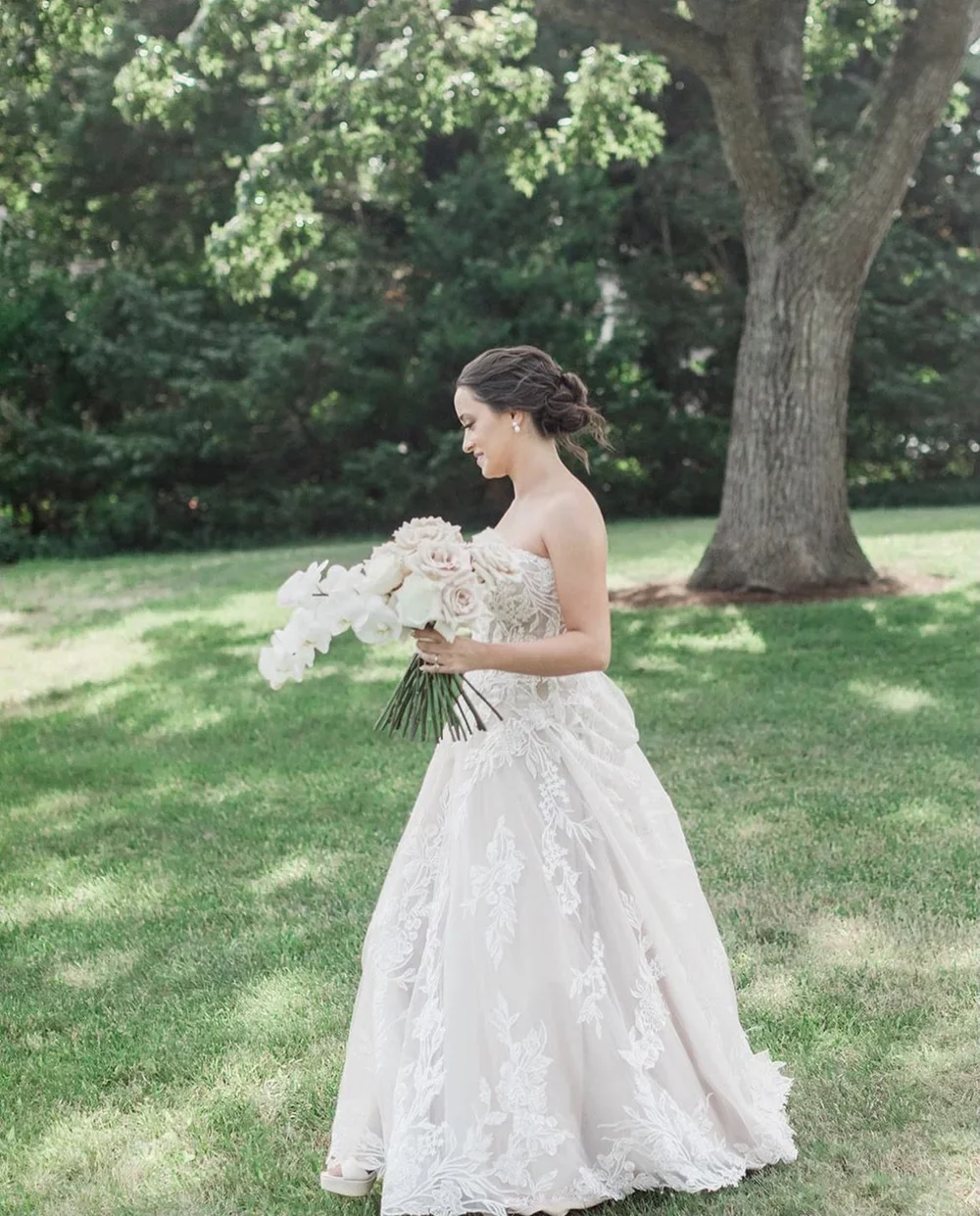 A bride in a strapless lace wedding gown holding a bouquet of white and blush roses standing on a grassy area under a large tree.