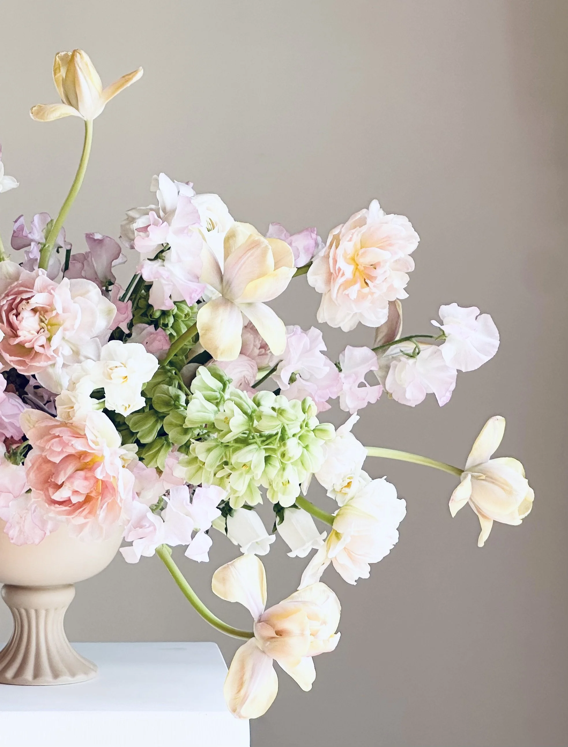 Arrangement of pastel-colored flowers including peonies, tulips, and hyacinths in a beige vase on a white surface against a neutral background.